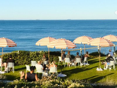 People in white chairs under striped umbrellas on a lawn, looking out over the ocean