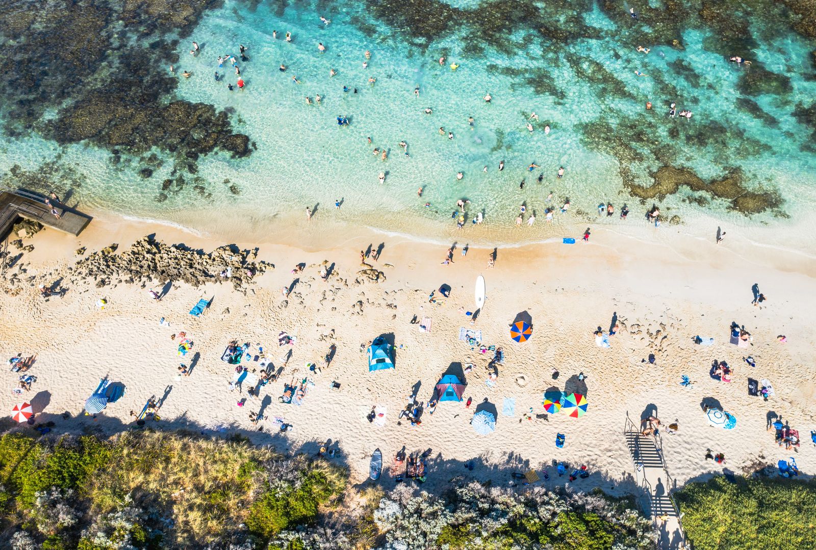 Mettams Pool from above: swimmers in a turquoise lagoon inside the reef, umbrellas on white sand, and the dune greenery behind