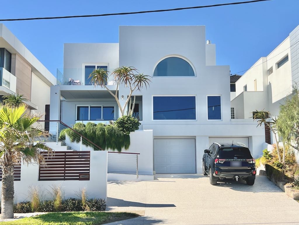 The white rendered house from West Coast Drive, with an arched window on the top floor and two garage doors