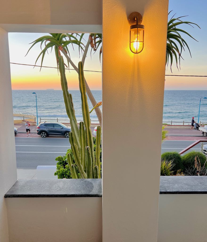 Sunset over the ocean seen between the white columns of the front porch, a wall lamp glowing